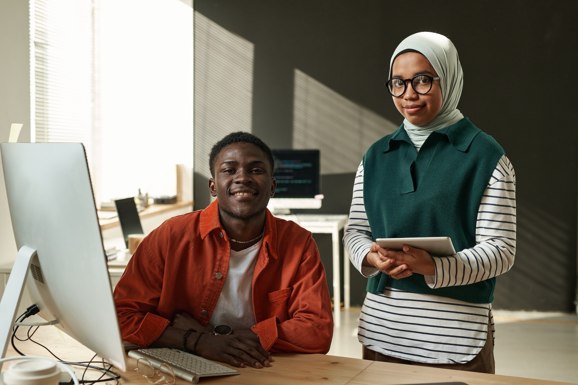 Young man sitting by desk and Muslim woman with tablet standing next to him